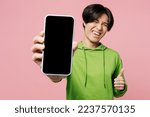 Young fun man of Asian ethnicity wear green hoody look camera hold in hand use close up mobile cell phone with blank screen workspace area show thumb up isolated on plain pastel light pink background