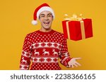 Small photo of Merry fun cool young man wear christmas sweater Santa hat posing hold toss up red present box with gift ribbon bow isolated on plain yellow background. Happy New Year 2023 celebration holiday concept