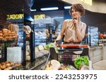 Small photo of Young minded happy woman 20s in casual clothes shopping at supermaket store with grocery cart buy choose pastry bread produce prop up chin inside hypermarket. People purchasing gastronomy food concept
