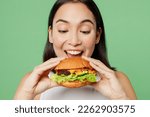Small photo of Close up young happy cheerful woman wear white clothes holding eating biting tasty burger isolated on plain pastel light green background. Proper nutrition healthy fast food unhealthy choice concept