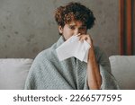 Young sad sick ill Indian man wapped in plaid sneezing have runny nose look camera sits on grey sofa couch stay at home hotel flat rest relax spend free spare time in living room indoor Lounge concept