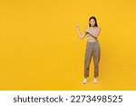 Full body fun young latin woman 30s she wear basic beige tank shirt point index finger aside indicate on workspace area copy space mock up isolated on plain yellow backround. People lifestyle concept