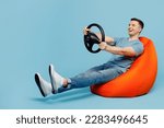 Full body side view young happy man wear casual t-shirt sit in bag chair hold steering wheel pretend driving car isolated on plain pastel light blue cyan background studio portrait. Lifestyle concept