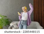 Young woman wear casual clothes headphones listen music dance raise up hand near grey sofa couch stay at home hotel flat rest relax spend free spare time in living room indoor People lounge concept