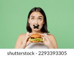 Small photo of Young wistful woman with mouth sealed with tape wear white clothes hold burger overhead area isolated on plain pastel light green background Proper nutrition healthy fast food unhealthy choice concept