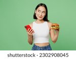 Small photo of Young minded fun woman wear white clothes hold eat burger use mobile cell phone look aside isolated on plain pastel light green background. Proper nutrition healthy fast food unhealthy choice concept