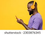 Side view young man of African American ethnicity wear casual clothes purple t-shirt headphones listen to music hold use mobile cell phone isolated on plain yellow background studio. Lifestyle concept