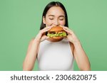 Young happy hungry fun cheerful woman wear white clothes holding eating biting tasty burger isolated on plain pastel light green background. Proper nutrition healthy fast food unhealthy choice concept