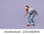 Full body side view young redhead bearded man he wears violet t-shirt casual clothes riding skateboard pennyboard isolated on plain pastel light purple background studio portrait. Lifestyle concept