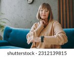 Elderly woman 50s year old wear casual clothes sits on blue sofa use look at smart watch put hand on neck check pulse stay at home flat rest relax spend free spare time in living room indoor grey wall