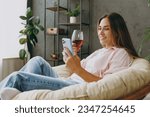 Side view young woman wear casual clothes sits in armchair hold wineglass drink wine use mobile cell phone stay home hotel flat rest relax spend free spare time in living room indoor Lounge concept
