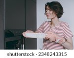 Small photo of Young housewife woman in casual clothes striped t-shirt puts casserole in baking dish in oven cooking sniff delicious scent food in light kitchen at home alone Healthy diet bakery lifestyle concept.