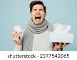 Small photo of Young sad ill sick man wear gray sweater scarf sneezing hold box of paper napkins cry sneeze isolated on plain blue background studio. Healthy lifestyle disease treatment cold season recovery concept