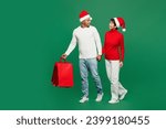 Full body young couple man woman wear red casual clothes Santa hat posing hold shopping package bag isolated on plain green background Black Friday sale buy day, Happy New Year 2024 Christmas concept