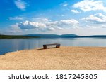 Small photo of One empty bench on deserted shore. Place for meditations, reflection and introspection. Amazing picturesque view of lake and mountains. Soft white clouds on blue sky.