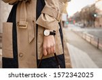 street style fashion details. close up, young fashion blogger wearing autumn trench coat and a white and golden black analog wrist watch. checking the time, holding a beautiful brown leather purse.
