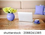 Open laptop, cup of coffee and very peri vase with beautiful yellow flowers on retro wood textured table in minimalistic living room with beige couch in background. Freelance workplace at home