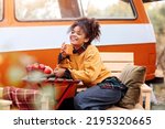 Young african american woman sitting at table near car in autumn forest and drinking hot tea, dreamy mixed race female relaxing around campsite, spending time in nature. Camping, travel and vanlife
