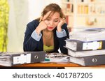 Business woman sitting by desk, paper files spread out, leaning head on hands looking overwhelmed and tired