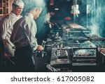 Food concept. Chef in white uniform monitors the degree of roasting and greases meat with oil in saucepan in interior of modern restaurant kitchen. Preparing traditional beef steak on barbecue oven.