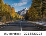 Amazing autumn view with an asphalt road in the mountains, first snow, forest and golden trees on the roadside against a background of blue sky and clouds. Altai, Russia