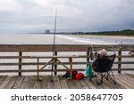 Small photo of MYRTLE BEACH, SC, USA - OCTOBER 8, 2021: An angler with two fishing rods relaxes in a folding chair on a fishing pier with an extensive beach view on a cloudy afternoon at Myrtle Beach State Park.