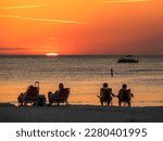 Small photo of Two mature couples in beach chairs watch the sun flatten out moments before it disappears over the Gulf of Mexico, while passengers on a small tour boat take in the view offshore. Southwest Florida.