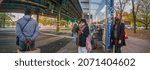Small photo of Passengers in coats stand and seat in masks waiting for MTA bus on a bus stop, White Plains Road and Pelham Parkway, New York, Very wide shoot panorama, Bronx, United States of America, 11.08.2021