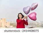 Smiling mature woman holding heart shape balloon while standing on building terrace