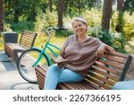 smiling modern old lady relaxing city park. Pensive senior gray haired woman casual sitting bench outdoors reading book. cycling forest park, bicycle, healthy active lifestyle after 50-60 years