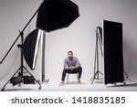 Professional photography studio showing behind the scenes lights. fashion handsome young man model at studio in the light flashes, sitting and looking at camera. indoor studio shot on grey background.
