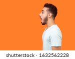 Side view portrait of surprised brunette man with beard in white t-shirt looking left with big eyes and open mouth, shocked by news, empty copy space for text. studio shot isolated, orange background
