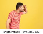 Profile portrait curious bearded man in red striped t-shirt trying to look far away holding hand on forehead, eye vision problems. Indoor studio shot isolated on yellow background