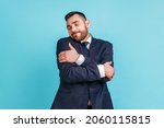 Small photo of I love myself! Portrait of narcissistic selfish young adult man in suit embracing herself, closed eyes and smiling with pleasure, complacency and egoism. Indoor studio shot isolated on blue background