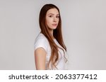 Small photo of Side view of portrait of strict bossy woman looking at camera, feels confident focused self-assured, expressing seriousness, , wearing white T-shirt. Indoor studio shot isolated on gray background.