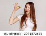 Portrait of beautiful woman holding golden bitcoin, showing btc coin and winking at camera with positive expression, wearing white T-shirt. Indoor studio shot isolated on gray background.