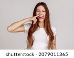 Small photo of Funny cheerful woman picking nose with stupid silly face, pulling out boogers, bad manners concept, misconduct, wearing white T-shirt. Indoor studio shot isolated on gray background.