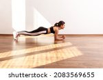Small photo of Side view of sportive woman practicing yoga, doing plank exercise on bent hands, training muscles, wearing black sports top and tights. Full length studio shot illuminated by sunlight from window.