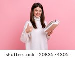 Small photo of Beautiful satisfied woman showing thumbs up gesture holding and reading book, likes genre and plot, wearing white casual style sweater. Indoor studio shot isolated on pink background.