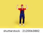 Full length portrait of optimistic woman worker standing and looking at camera with raised arms, showing her power, wearing overalls and red cap. Indoor studio shot isolated on yellow background.