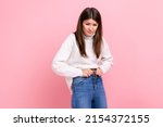 Small photo of Portrait of unhappy sad young adult woman gaining weight, cant wearing her jeans, being overweight, wearing white casual style sweater. Indoor studio shot isolated on pink background.