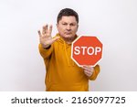 Concentrated serious man looking angrily showing stop gesture, holding road traffic sign, warning of ban, forbidden access, wearing urban style hoodie. Indoor studio shot isolated on white background.