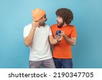 Small photo of Portrait of two excited amazed young adult hipster men standing with cell phone and celebrating success, clenched fists, looking at each other. Indoor studio shot isolated on blue background.