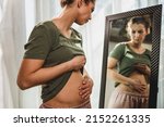 Small photo of Frown young woman standing in front of a mirror and holding hands on her bloating stomach.