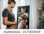 Small photo of Frown young woman standing in front of a mirror and holding hands on her bloating stomach.