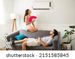 Sad woman feeling hot and trying to cool down sitting below the air in the ac unit with her partner 