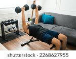 Strong fit man in his 20s with a home gym doing bench press exercises with dumbbell weights