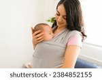 Small photo of Smiling happy mother feeling a lot of love for her infant child while using a baby sling carrier around the house
