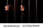 hands of a prisoner behind prison bars on black background

