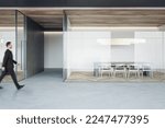 Businessman walking on concrete floor by stylish conference rooms with minimalistic style furniture on wooden parquet floor behind glass partitions in spacious office hall
